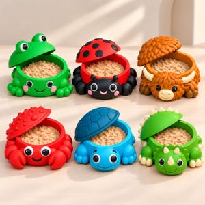 Pode incluir: Um conjunto de seis recipientes em forma de animal com tampas, cada um preenchido com uma substância granular bege. A coleção inclui um sapo verde, uma joaninha vermelha, uma vaca castanha, um caranguejo vermelho, uma tartaruga azul e um triceratops verde.