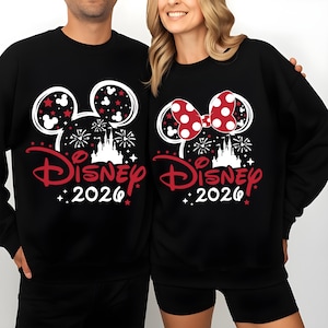 May include: Black sweatshirts with Disney-themed designs. One features Mickey Mouse ears with red stars, the other a red polka-dot bow. Both have a castle graphic, fireworks, and the text "Disney 2026" in red.