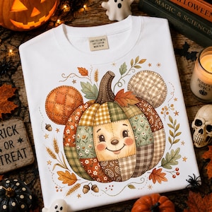 Puede incluir: Una camiseta blanca con un gráfico central de una calabaza de patchwork con cara sonriente y dos orejas acolchadas circulares. La calabaza está rodeada de hojas de otoño, tallos de trigo y pequeñas estrellas. La etiqueta interior dice UGNT YELLOW TEE.