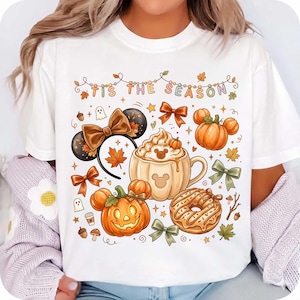 May include: A white t-shirt featuring a colorful graphic print with the text 'TIS THE SEASON'. The design includes a pumpkin-shaped mug with whipped cream, a jack-o'-lantern, a donut, mouse ears with a bow, autumn leaves, and bows.