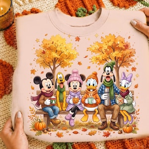 May include: A light peach-colored sweatshirt featuring a graphic print of Mickey Mouse, Pluto, Minnie Mouse, Donald Duck, Goofy, and Daisy Duck sitting on a wooden bench. The characters are wearing autumn clothing and holding mugs amidst fall trees.