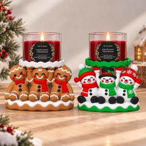 May include: Two candle holders featuring festive figures. The left holder has three gingerbread people in red scarves supporting a glass candle with a red wax Mahogany Teakwood label. The right holder has three snowmen in winter hats and scarves supporting an identical candle.