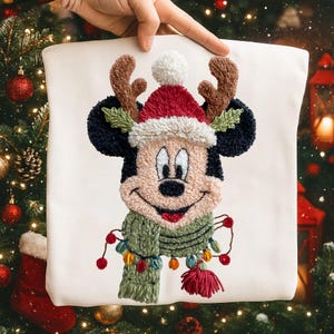 May include: A cream-colored decorative pillow featuring a festive Mickey Mouse design. Mickey is dressed in a Santa hat, reindeer antlers, and a green scarf adorned with Christmas lights. The design is made with textured embroidery.