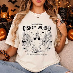 May include: A cream-colored t-shirt featuring a black graphic print of a character dressed as a ghost walking down a street toward a castle. The text reads FLORIDA EST. 1971 DISNEY WORLD and The Scariest Place on Earth with bats and ghosts.