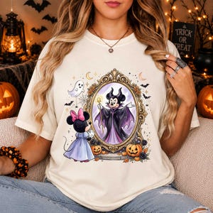 May include: A cream-colored t-shirt featuring a graphic of a character in a purple dress looking into an ornate gold mirror. Inside the mirror, a character dressed as a villain with a staff is reflected. The design includes pumpkins, bats, and a ghost.
