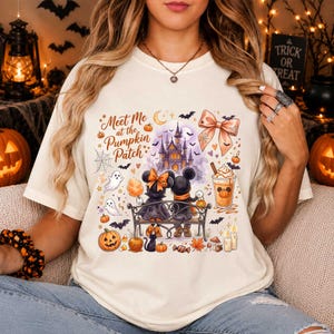 May include: A cream-colored t-shirt featuring a graphic print of two cartoon mice sitting on a bench in front of a castle. The design includes the text Meet Me at the Pumpkin Patch, a bow, ghosts, pumpkins, bats, a drink, and candles.