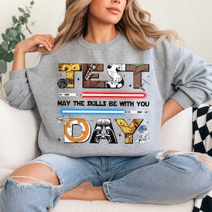 May include: A gray sweatshirt with the words "TEST DAY" in a Star Wars-themed design. The text "MAY THE SKILLS BE WITH YOU" is between two lightsabers. The design includes characters like C-3PO, Darth Vader, and R2-D2.