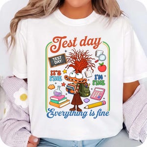 May include: White t-shirt with a colorful graphic featuring a cartoon character with books and a backpack. The design includes the text "Test day", "It's fine", "I'm fine", and "Everything is fine", along with school-themed illustrations.