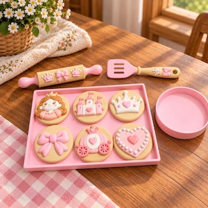 May include: A toy baking set featuring a beige rolling pin with pink crowns and bows, a pink spatula, and a square pink tray holding six round beige cookies decorated with pink and white princess-themed motifs including a carriage and heart.