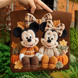 May include: A brown decorative pillow featuring Mickey and Minnie Mouse characters in a fall theme. The characters are wearing sweaters and are surrounded by pumpkins, leaves, and plaid bows. The pillow is held by a hand.