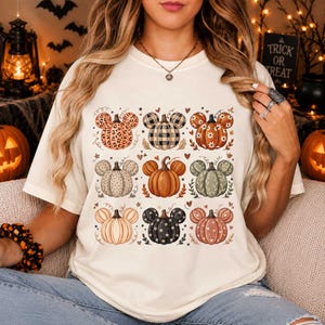 Può includere: Una t-shirt color crema con una griglia di nove illustrazioni di zucche, ognuna con forme a orecchie di topo. Le zucche presentano vari motivi tra cui stampa leopardata, plaid, fiori e colori solidi, accentuati da piccoli cuori e foglie.