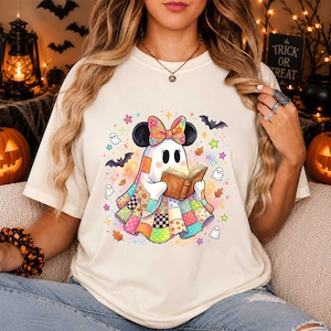 Puede incluir: Una camiseta de color crema con un gráfico de un fantasma que lleva una manta de patchwork colorida y un lazo floral, leyendo un libro abierto. El diseño incluye pequeños fantasmas, murciélagos y estrellas de varios colores.