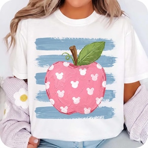 May include: A white short-sleeve t-shirt featuring a central graphic of a pink apple with a green leaf and brown stem. The apple is decorated with small white mouse-ear silhouettes and is layered over horizontal blue brushstroke stripes.