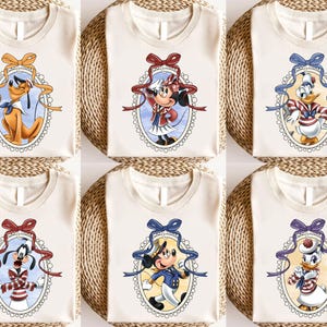 以下が含まれることがあります： 異なる漫画のキャラクターデザインが施された白いTシャツ6枚。各デザインは、青、赤、または黄色のリボンが付いた円形のフレームの中にキャラクターが描かれています。キャラクターはミッキーマウス、ミニーマウス、ドナルドダック、グーフィー、プルート、デイジーダックです。