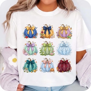 May include: A white short-sleeved t-shirt featuring a graphic print of nine colourful pumpkins arranged in a grid. Each pumpkin is decorated with a large bow and various thematic elements, including flowers, books, seashells, a lamp, and a glass slipper.