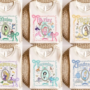 以下が含まれることがあります： さまざまなディズニーのプリンセスデザインが施された、クリーム色のTシャツ。各シャツには、関連するイラストとリボンが付いた装飾フレームにプリンセスの名前が表示されています。プリンセスには、ムーラン、アリエル、ベル、ラプンツェル、ジャスミン、シンデレラが含まれます。