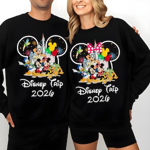 Op de afbeelding: Zwarte sweatshirts met een Disney Trip 2026-ontwerp. Het ontwerp bevat Mickey Mouse, Minnie Mouse, Donald Duck, Goofy en andere personages, een kasteel en vuurwerk. Eén sweatshirt heeft een roze strik, de andere een gele strik.