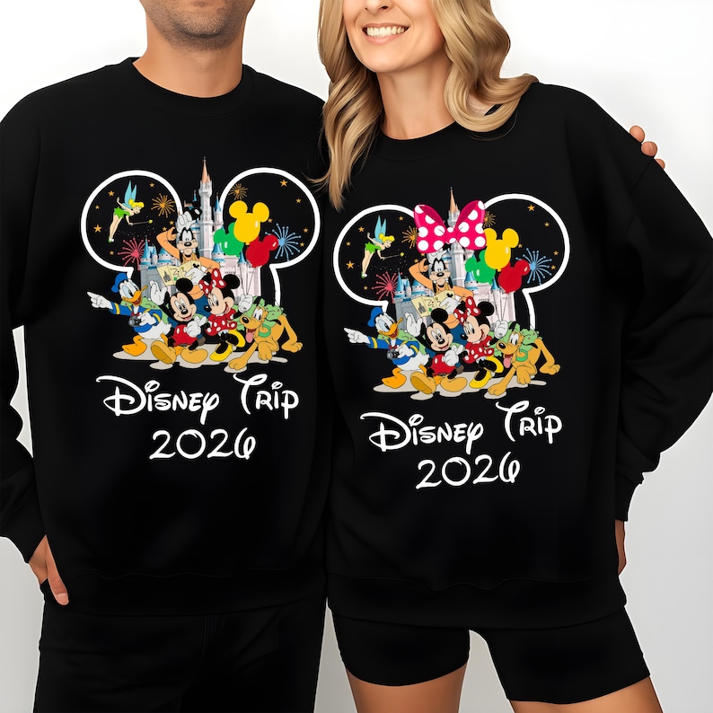 May include: Black sweatshirts featuring a Disney Trip 2026 design. The design includes Mickey Mouse, Minnie Mouse, Donald Duck, Goofy, and other characters, along with a castle and fireworks. One sweatshirt has a pink bow, and the other has a yellow bow.