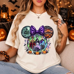 May include: A cream-colored t-shirt featuring a graphic of Disney villains arranged in a Mickey Mouse ear shape. The design includes a purple bow, a haunted castle, a volcano, and several pumpkins. The characters depicted are Jafar, Maleficent, the Evil Queen, Ursula, and Hades.