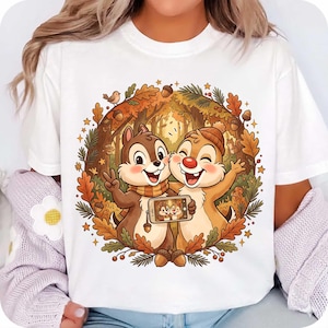 May include: A white t-shirt featuring a circular graphic of two cartoon chipmunks in an autumn forest setting. The chipmunks are taking a selfie with a smartphone, surrounded by oak leaves, acorns, and small stars in shades of brown and orange.