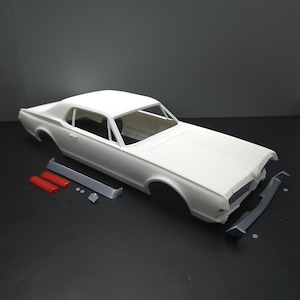 May include: A white model car kit, featuring a classic coupe design. The kit includes the car body, bumpers, and other components. The model car is unassembled, with parts in white, red, and gray.