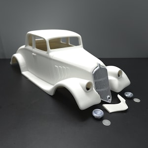 May include: A white model car body with detached parts, including fenders, a grill, and wheels. The classic car design features a coupe body style with a detailed front end and a streamlined roof. The parts are arranged on a dark surface.
