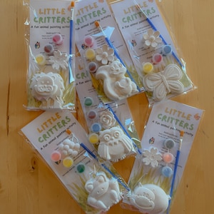 Könnte beinhalten: Sechs Little Critters Mal-Sets in durchsichtigen Plastiktüten. Jedes Set enthält eine weiße Tier- oder Blumenfigur aus Gips, einen kleinen blauen Pinsel und mehrere kleine, runde Farbtöpfchen mit verschiedenen Farben zum Bemalen.