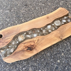 May include: Wooden serving board with a natural edge and a river of seashells and resin. The board is light brown with a dark brown resin river filled with various seashells. It has a hole for hanging.