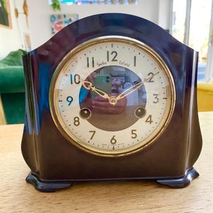 May include: A dark brown mantel clock with a cream-coloured circular face, gold-toned hands, and a gold-toned bezel. The clock face features black Arabic numerals and the text Smiths Enfield and Made in Great Britain. It has a curved top.