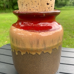 May include: A ceramic jar with a cork lid. The jar has a red, brown, and yellow glaze. The jar is sitting on a wooden table.