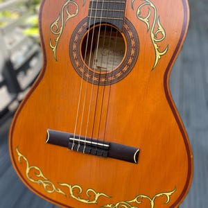 May include: A brown acoustic guitar with a dark wood bridge and fretboard. The body features a decorative dark brown border and yellow vine-like patterns painted on the soundboard. A circular sound hole is surrounded by a patterned rosette.
