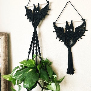 May include: Two black macrame bat wall hangings with white wooden dowels. The larger bat is hanging over a plant in a white pot.