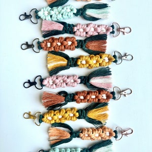 May include: A set of eight colorful macrame keychains with tassels. Each keychain features a different color combination of green, brown, pink, yellow, and white yarn.