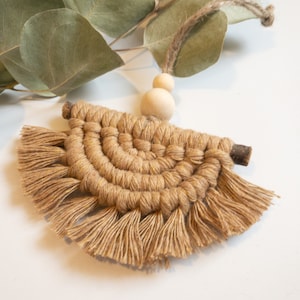 May include: A small, beige macrame wall hanging with a wooden bead at the top. The macrame is in the shape of a half circle and has fringe around the bottom.