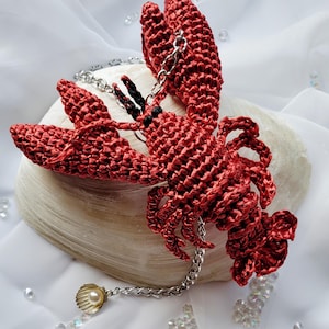 以下が含まれることがあります： 銀色のチェーンと貝殻のペンダントに真珠が入った、赤い毛糸で編まれたロブスターのチャームです。