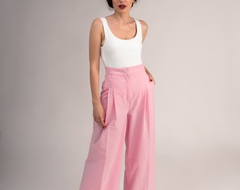 May include: A woman wearing a white tank top and pink wide-leg pants. She is standing in front of a white background.