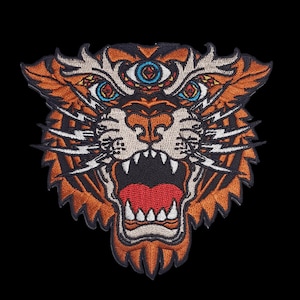May include: Embroidered patch featuring a detailed tiger head with an open mouth, sharp teeth, and a third eye. The design incorporates orange, black, white, and red colors, with intricate stitching details. The patch is likely for clothing or accessories.