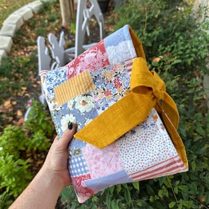 May include: A patchwork fabric clutch with a mustard yellow bow closure. The clutch features a variety of floral and patterned fabrics in shades of red, blue, pink, and yellow. The bag is quilted and appears to be handmade.