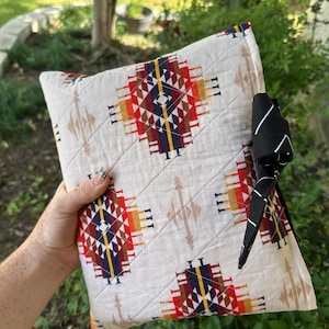 May include: A quilted fabric pouch featuring a cream-colored base with a repeating geometric pattern in red, orange, yellow, and dark blue. The pouch is secured with a black fabric tie featuring thin white lines, creating a functional closure.