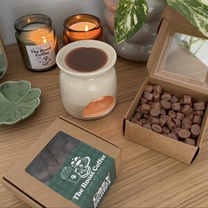 Op de afbeelding: Een op koffie ge&iuml;nspireerde geurcompositie voor in huis. Het bevat een keramische waxbrander met een bruine waxmelt, twee brandende kaarsen en een doos met koffieboonvormige waxmelts. De branding van The Roost Coffee is zichtbaar.