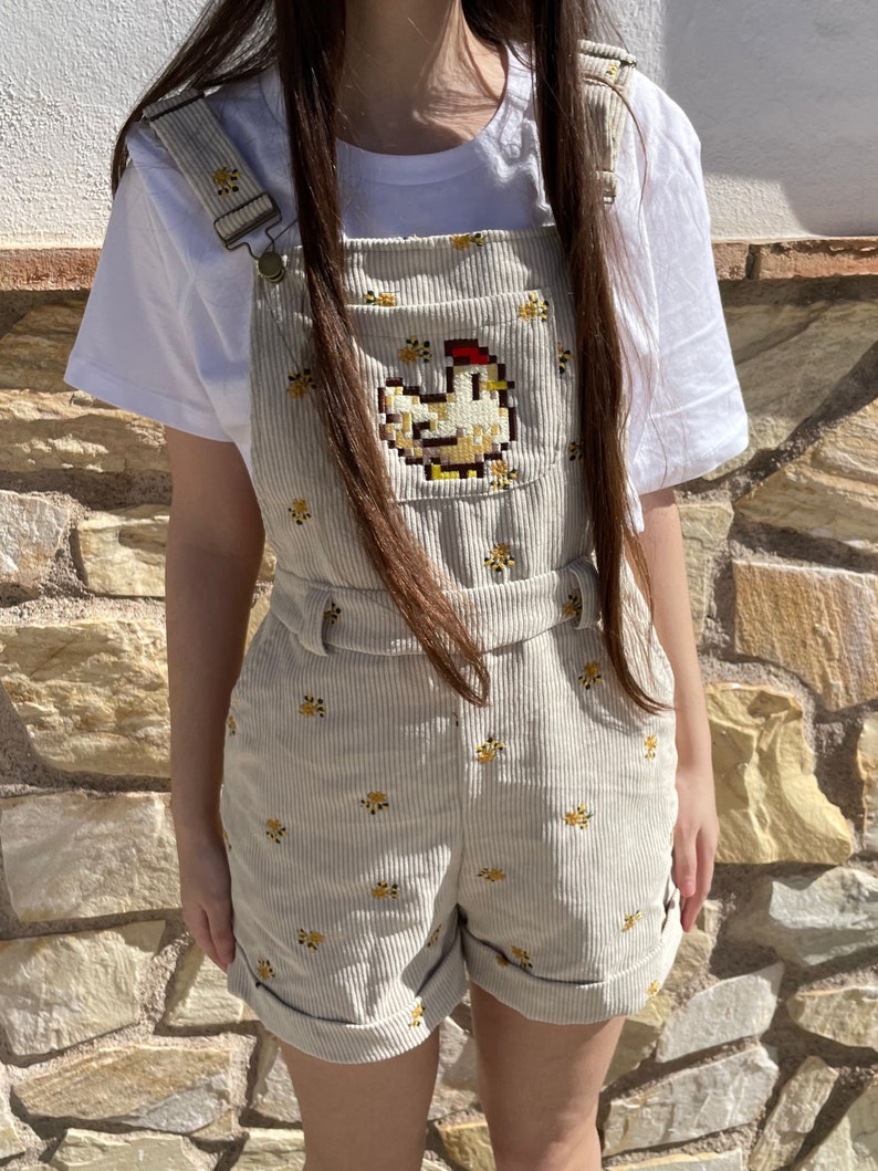 May include: Beige corduroy overall shorts featuring a pixelated chicken design on the front. The shorts have a repeating floral pattern and adjustable straps. A white t-shirt is worn underneath.