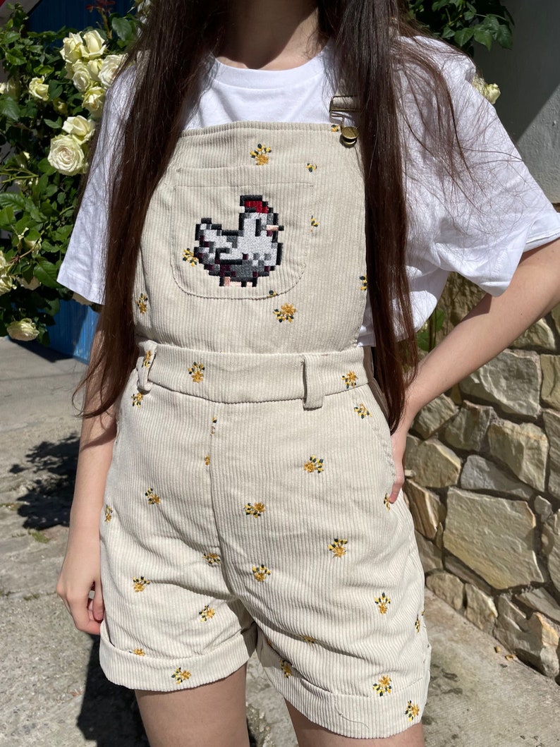 May include: Beige corduroy overall shorts with a pixelated chicken design on the bib. The shorts feature small yellow floral embroidery and a belt. The overall straps have metal hardware.