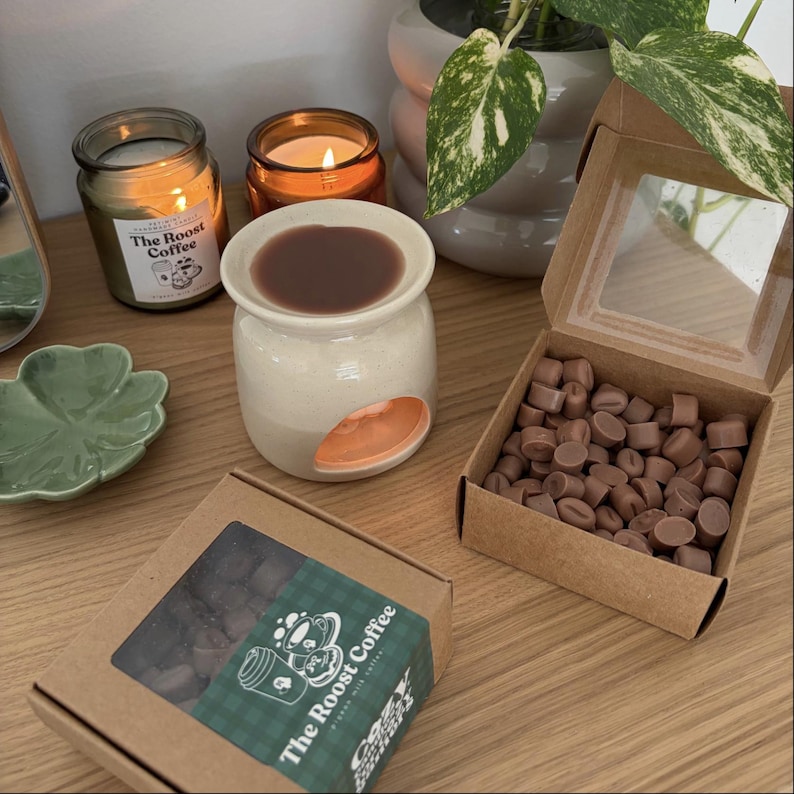 Op de afbeelding: Een op koffie ge&iuml;nspireerde geurcompositie voor in huis. Het bevat een keramische waxbrander met een bruine waxmelt, twee brandende kaarsen en een doos met koffieboonvormige waxmelts. De branding van The Roost Coffee is zichtbaar.