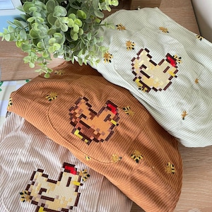 Könnte beinhalten: Drei Cordhüte in Creme, Braun und Hellgrün, jeweils mit einem gestickten pixeligen Hühnerdesign. Kleine gestickte Bienenakzente verzieren die Hüte und suggerieren ein Bauernhof- oder Landthema.