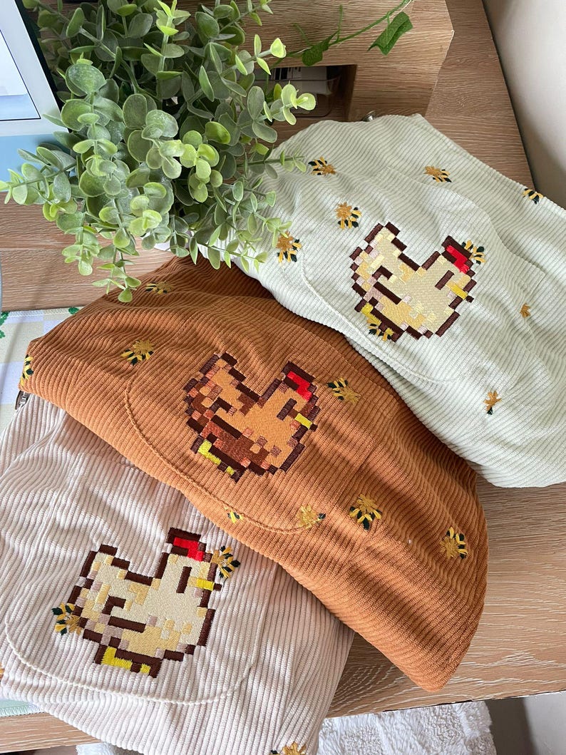 May include: Three corduroy hats in cream, brown, and light green, each with an embroidered pixelated chicken design. Small embroidered bee accents decorate the hats, giving a farm or country feel.