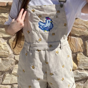May include: Beige corduroy overall shorts featuring a blue pixelated chicken design on the front pocket. The shorts are decorated with small yellow floral embroidery. The overall straps have metal clasps and a matching belt.