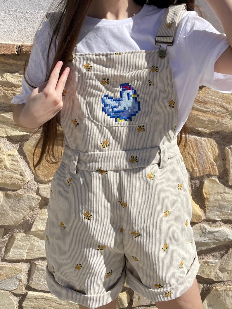 May include: Beige corduroy overall shorts featuring a blue pixelated chicken design on the front pocket. The shorts are decorated with small yellow floral embroidery. The overall straps have metal clasps and a matching belt.