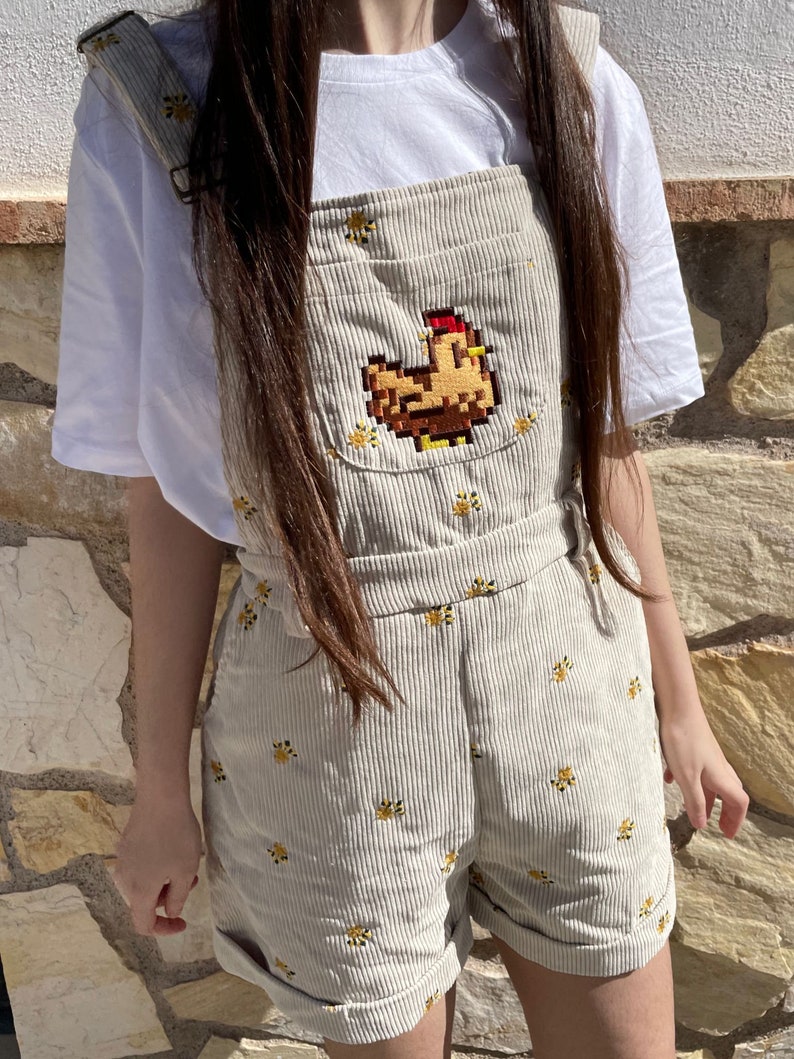 May include: Beige corduroy dungarees with a pixelated chicken design on the front pocket. The dungarees feature yellow floral embroidery and adjustable straps. The shorts are cuffed at the hem. A white t-shirt is worn underneath.