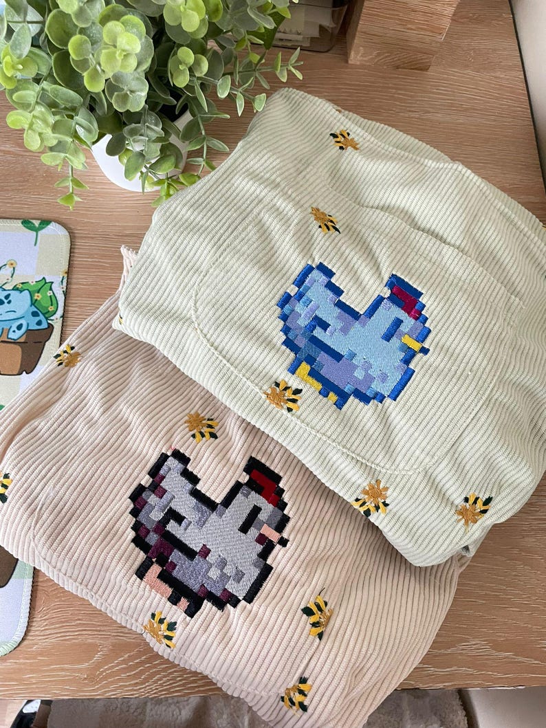 May include: Two corduroy items, one light beige and one light green, are stacked. Each features a pixelated chicken design and small embroidered bees. The items are on a wooden surface.