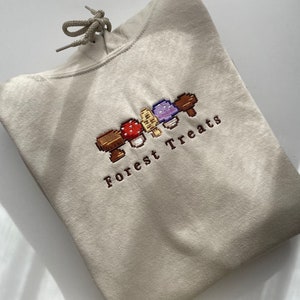 Może przedstawiać: Beżowa bluza z kapturem ze sznurkiem i haftowanym wzorem "Forest Treats". Wz&oacute;r przedstawia pikselowane grzyby w kolorze czerwonym, brązowym, fioletowym i ż&oacute;łtym. Bluza z kapturem jest złożona, pokazując prz&oacute;d z haftem.
